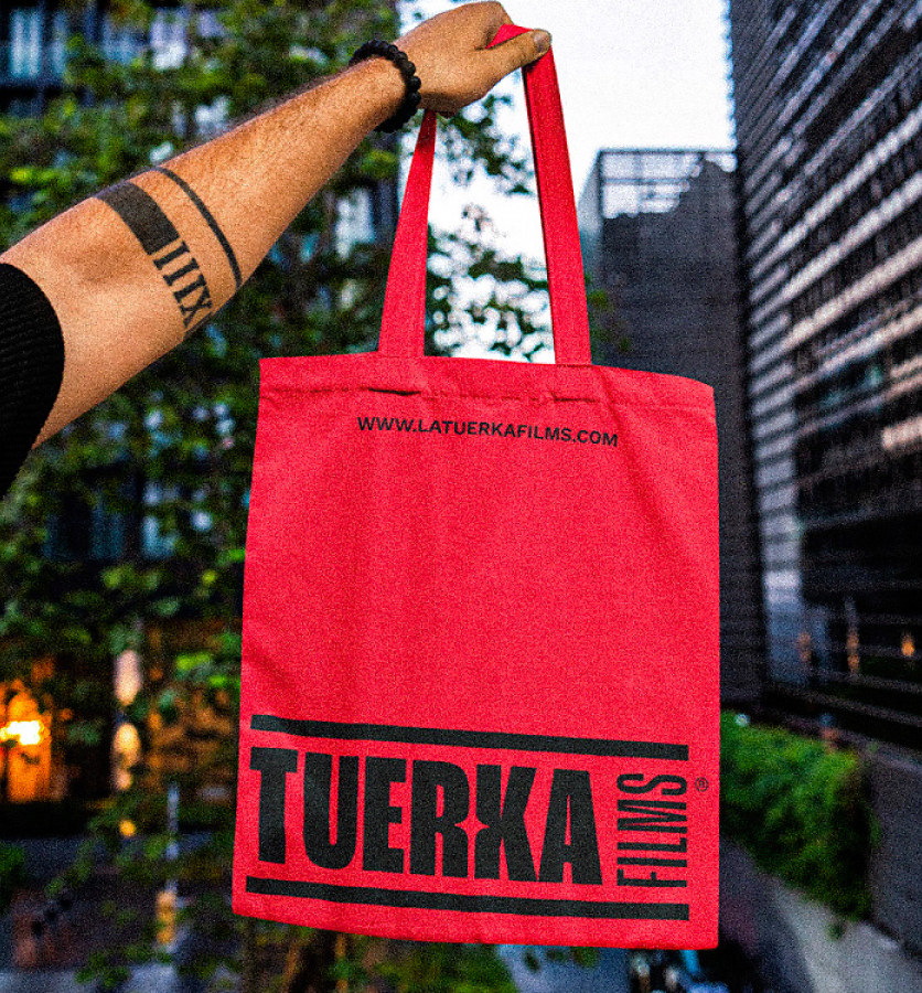 Joven, con tatuaje, mostrando una totebag roja de La Tuerka Films