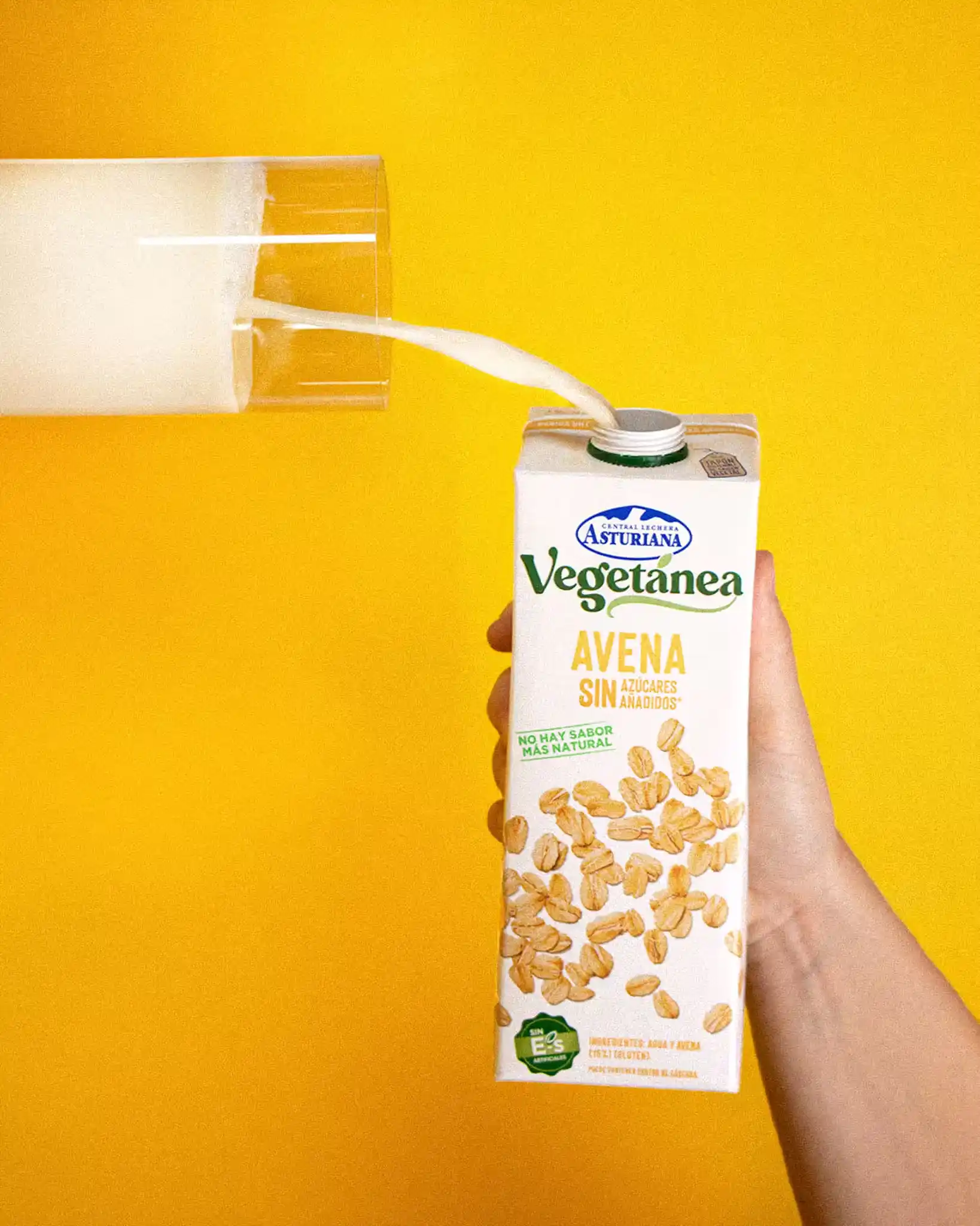 Mano sujetando una caja de leche de avena vegetánea que está llenando un vaso venciendo a la gravedad